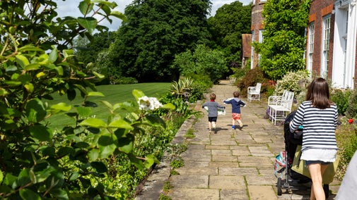 A paved garden path runs alongside a brick house with white doors and windows, bordered by lush plants and flowers. Two children run ahead along the path while an adult pushes a stroller behind them, with a large green lawn and tall trees stretching out to the left in bright summer sunshine.